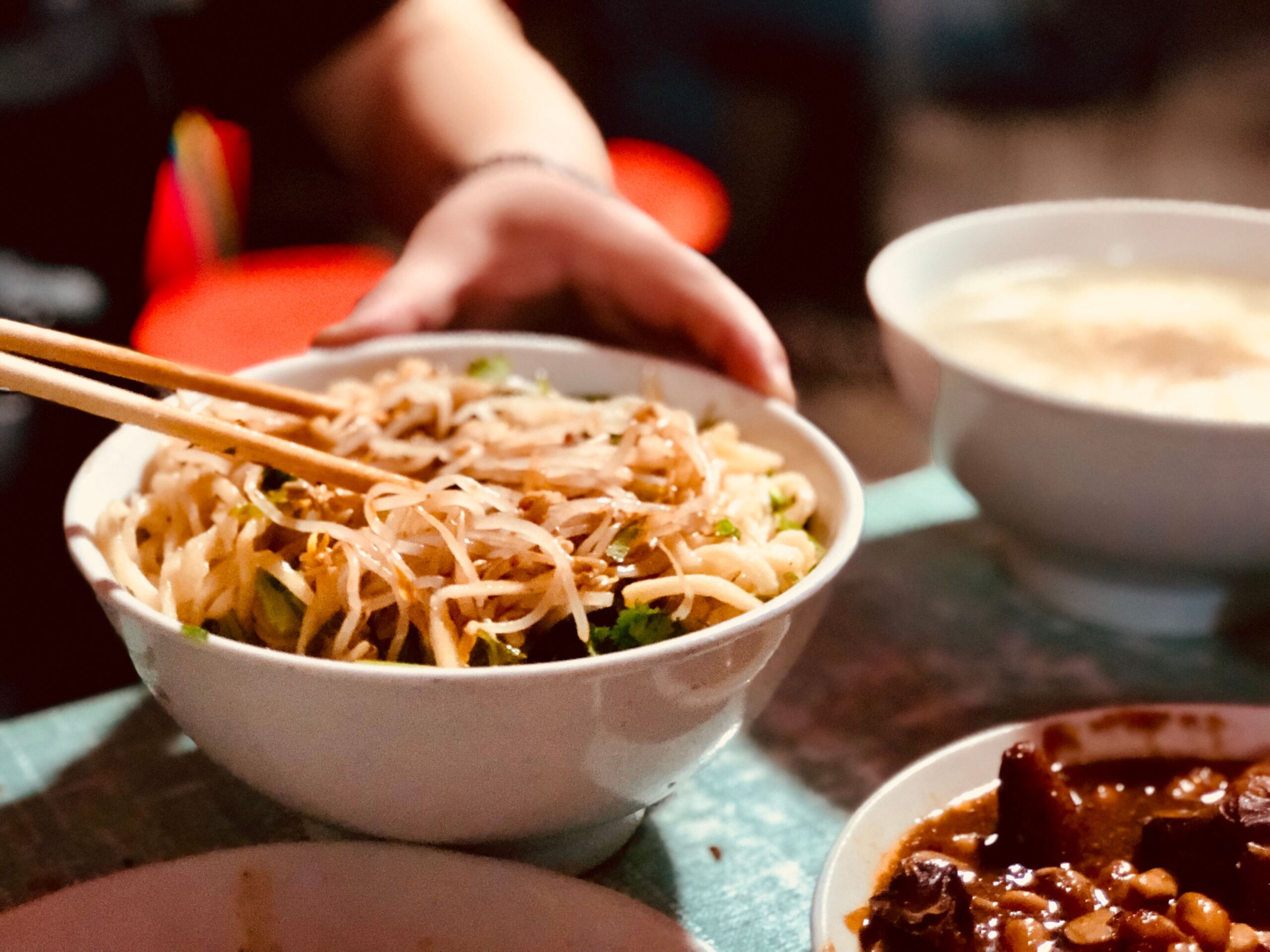 Un plato de fideos picantes, con una manos de fondoa agrrando unos palillos